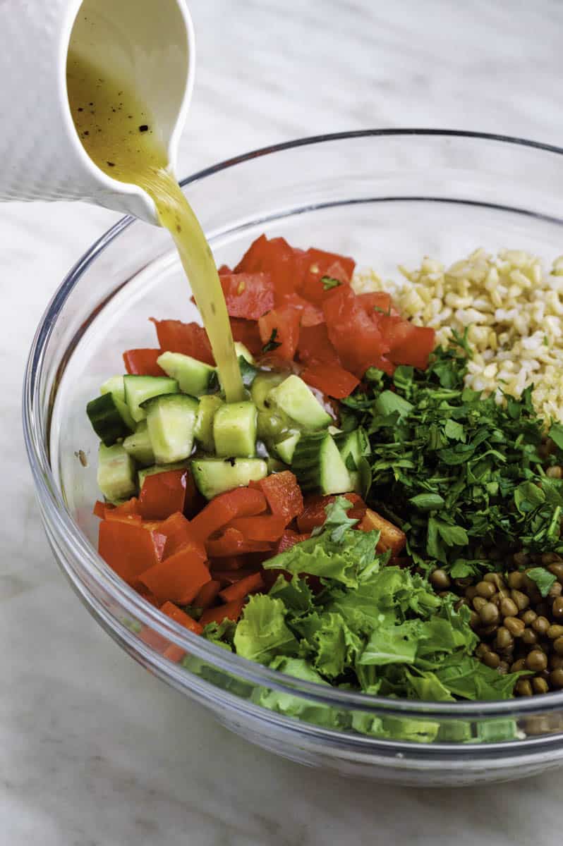 Pouring the salad dressing from a white jug into a glass bowl filled with vegetables.