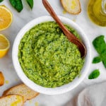 Cashew pesto served in a white bowl with a wooden spoon in it. Sliced baguette, basil leaves and halved lemon placed next to the bowl.