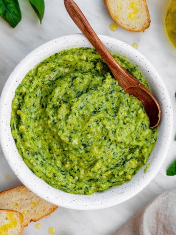 Cashew pesto served in a white bowl with a wooden spoon in it. Sliced baguette, basil leaves and halved lemon placed next to the bowl.