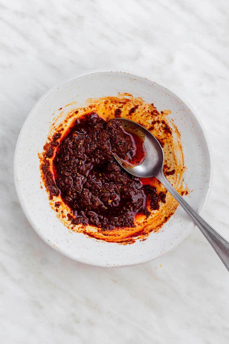 A paste made from spices, soy sauce, and oil in a white bowl with a spoon in it.