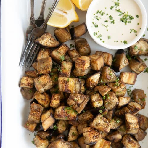 overhead of roasted eggplant cubes with three forks, lemon wedges, and side of dipping sauce.