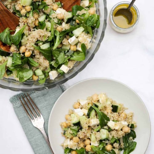 A bowl with a wooden spoon filled with a salad and a white in front with a serving on it.