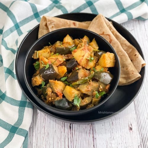 eggplant potato curry in a black bowl on a black serving dish with side of roti.