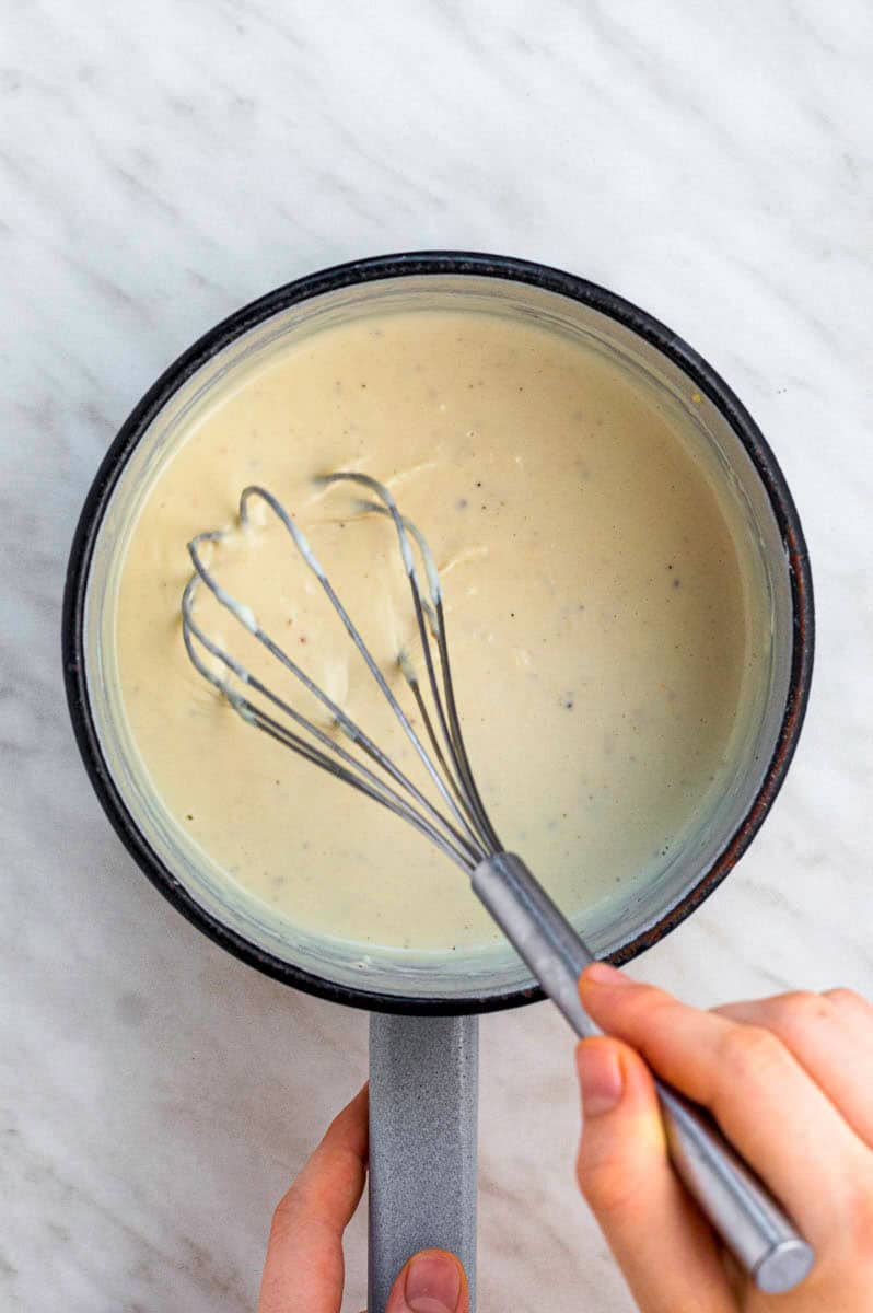 Mixing white sauce with a stainless-steel whisk from a white pot.