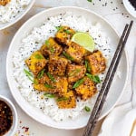 A plate filled with cooked white rice topped with honey garlic tofu. Everything is garnished with black sesame seeds and chopped scallions.