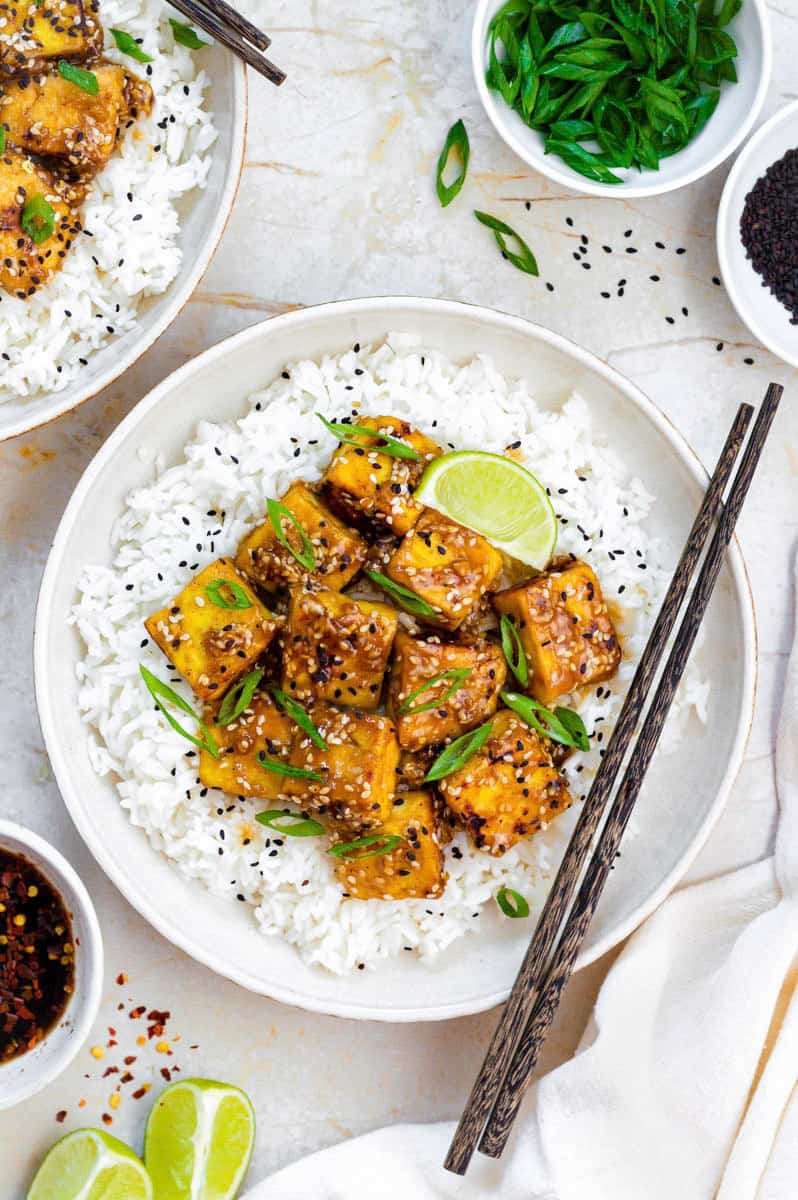 A plate filled with cooked white rice topped with honey garlic tofu. Everything is garnished with black sesame seeds and chopped scallions.