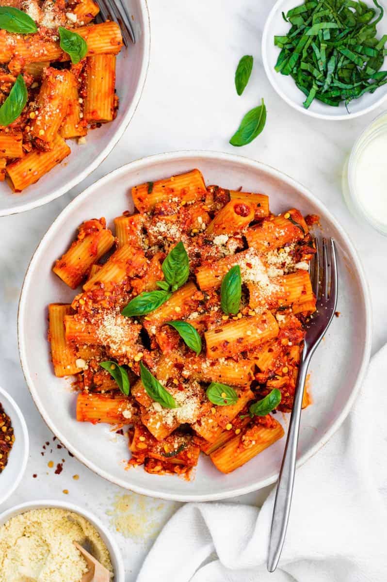 A plate filled with tofu pasta topped with vegan parmesan and basil. A fork placed on the right side of the plate.