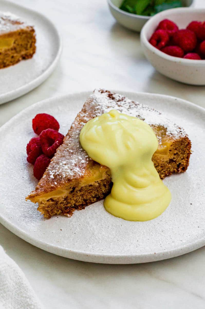 A piece of apple cake serve in a small white plate and topped with powdered sugar and pastry cream.