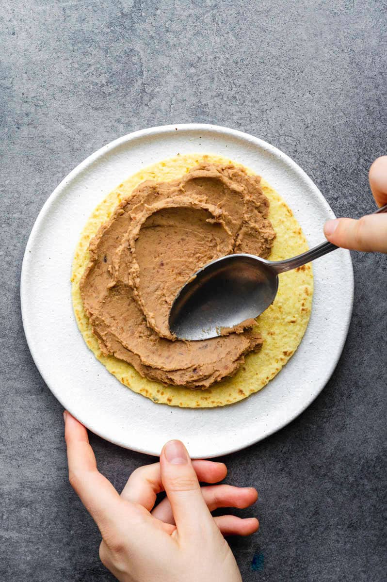 A corn tortilla placed on a white plate. A hand spreading refried beans on the tortilla using a silver spoon.