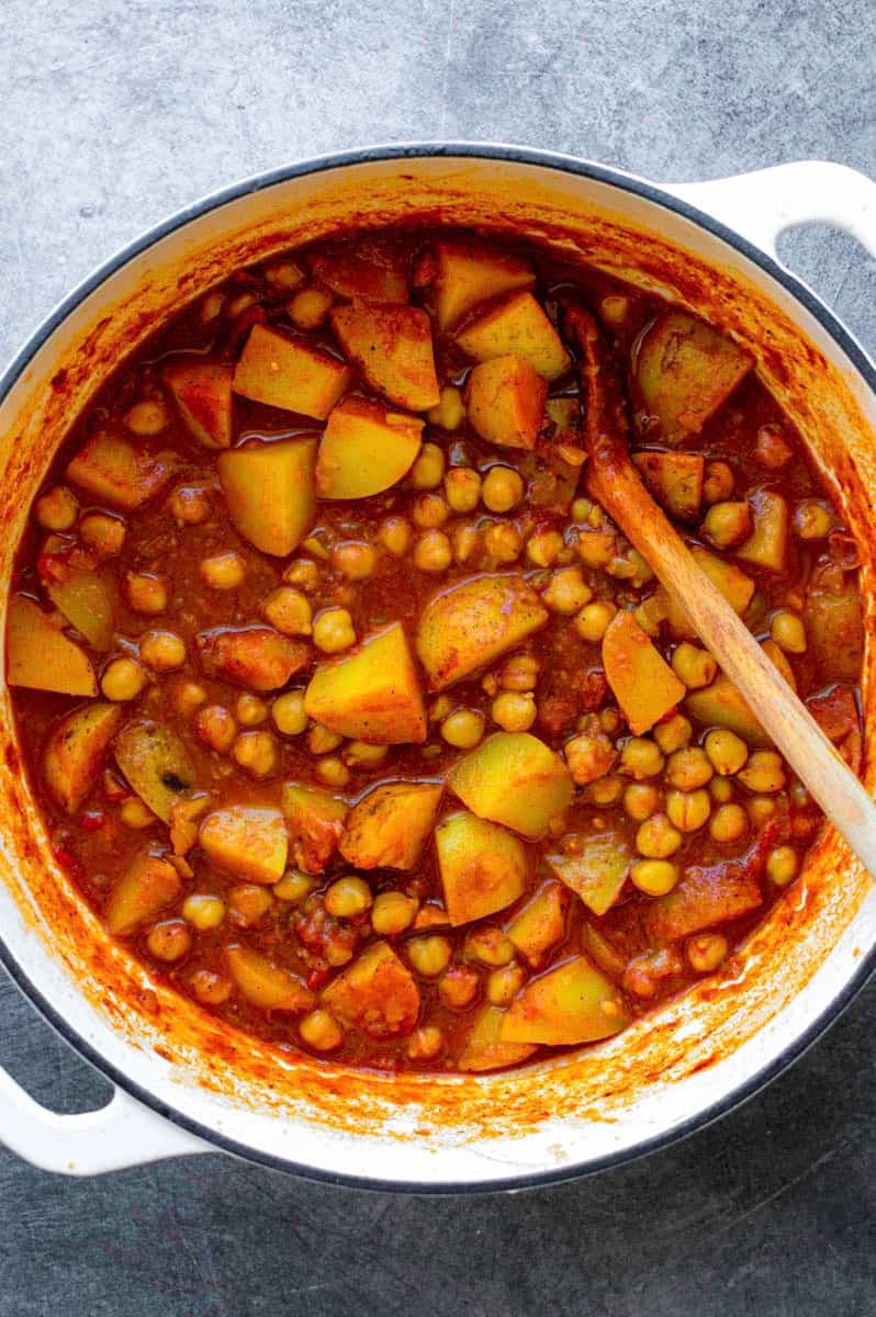 Chickpea and potato curry in a white Dutch oven with a wooden spoon in it.