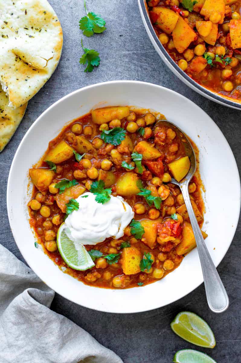 A white bowl filled with chickpea and potato curry topped with sour cream, fresh cilantro, and a lime wedge placed on the left side of the bowl.