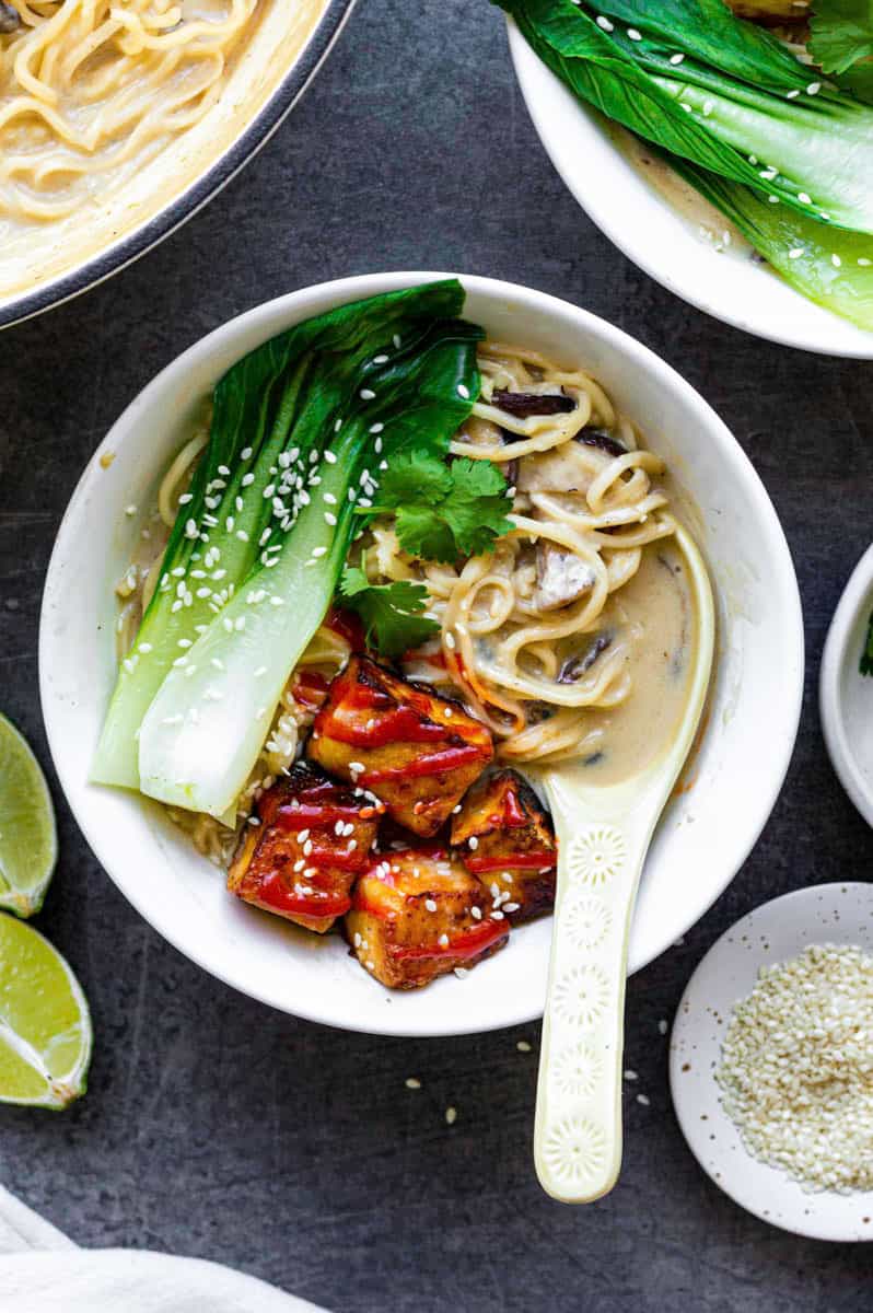 A white bowl filled with vegan ramen, topped with fried tofu, bok choy, and sesame seeds. A white soup spoon placed on the right side of the bowl.