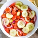 overhead of cucumber tomato salad in white bowl with wooden spoon to the right.