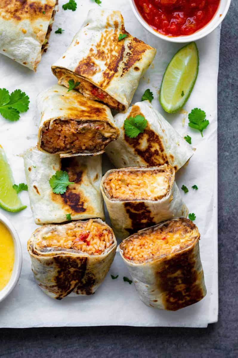 Halved burritos served on a white board layered with parchment paper, topped with fresh cilantro. Lime wedges placed on both sides of the burritos.