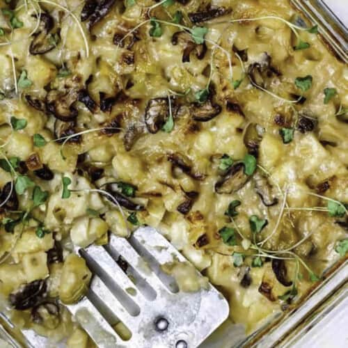 Vegan potato bake in a glass casserole dish, topped with microgreens and with a metal spatula dug in it.