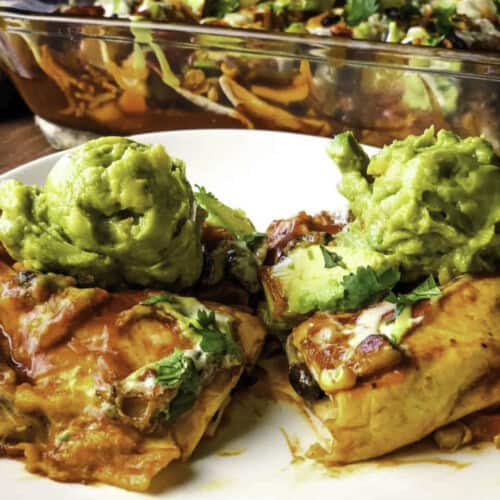 Black bean enchiladas topped with guacamole, served on white plate with casserole dish in background.