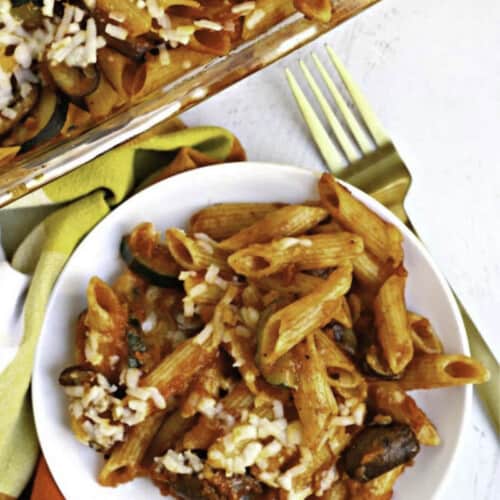 Plate of baked penne pasta with casserole in the background.