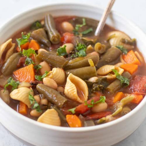 close-up of minestrone soup in a white bowl with a spoon in it.