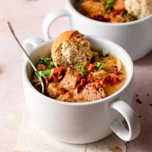 Vegan zuppa toscana served in a small white bowl with two handles on each side. A piece of bread dipped in the soup.