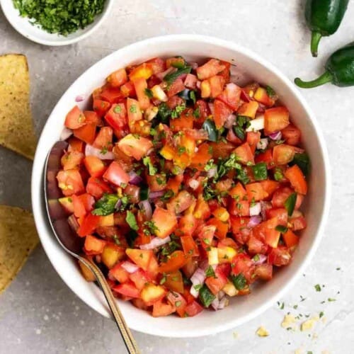 A bowl of fresh tomato salsa served with spoon with chips, jalapenos, and chopped cilantro beside.